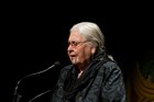 Woman with gray hair speaking into a microphone in front of black background