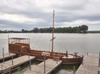 A historic flat bottomed wooden boat with a mast, sitting on a river at a dock.