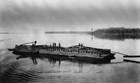Black and white image of a flat wooden boat on a river.