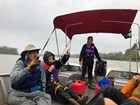 Young person operating the engine of an open boat in the rain. Two people point into the distance.
