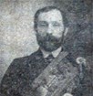 Newspaper photograph of man with dark hair and beard wearing suit and regalia.