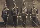 Four men in a line wearing feathered hats, regalia, and holding swords.