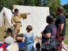 Person in early 1800s explorer costume speaks with family, points to the right. Canvas tent behind.