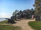 A historic building made of timber stands on the ocean shoreline fencing and a large cannon.