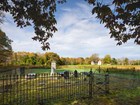 Gravestones are surrounded by a black iron fence, a brick farmhouse sits in a field grass and trees.