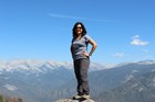 Woman smiles as she stands atop granite rock, spikey mountain ridge and blue skies in background.