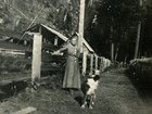 Older woman in plain dress leans against wooden fence at family homestead with her dog in foreground