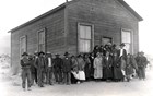Large group of Owens Valley Paiute in front of one-room wooden building in shrubby landscape.