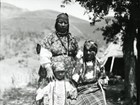 Indigenous woman in dress and headband stands with arms around an older seated woman and young child