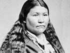 Native American woman with long hair, stripped shawl and beaded necklace, gazes at camera.
