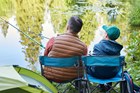 An adult and child sitting in camp chair and fishing by a lake.