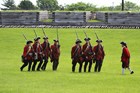 Several men in varying red uniforms walk in formation outside the fort wall. 