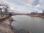 A low flow river with high dirt banks and leafless trees and brown grass and shrubs on the banks.
