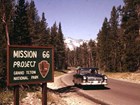 A 1960s-era car drives past an NPS sign reading 