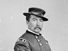 An 1864 portrait photo shows a mustached man seated, hand-in-waistcoat, in an army uniform.