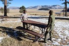 Two maintenance workers move a picnic table in a grassy, open area.