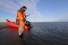 A scientist looks at science gear while standing near the shore with a packraft in the background. 