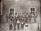 Group of 17 men standing in front of a wood frame building. 
