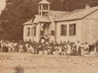 Sepia-toned image of students standing outside of a small, white wooden school building.