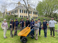 Ten people holding garden tools and wheelbarrows stand in front of lilacs and a two-story mansion.