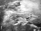 Black and white photo of two planes flying above clouds with ground visible below. 