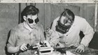 Young man wearing goggles sits at a workbench to weld. Another man inspects his work.