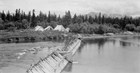 1940s photo of a wooden weir across the water. A group of tents stands on the opposite shore.