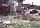 A person kneels by a hole behind a pile of dirt during an excavation behind a log structure.
