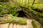 Mossy stones and a metal railing line a staircase leading to a dark cave opening in a wooded area.
