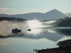 Fishing boats float in still water, surrounded by bright mist with mountains in the background.