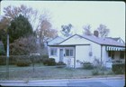 Color photo of a small 1960s era white house with trees, lawn, and street visible