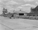 Black and white of an air strip with an airplane on the runway and a line of soldiers to the right