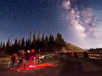 A group of people with red lights and telescopes under a starry night sky with the Milky Way visible
