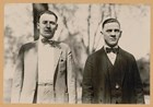 Two men in suits and hats standing next to one another, shown from the waist up.