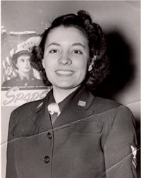 Black and white photo of woman in military uniform smiling in front of SPARS recruitment poster