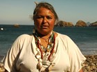 Indigenous woman in shell necklace stands  on beach, ocean and islands in background.