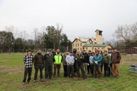A group of people in hats, coats, and work clothes stand in an orchard area in front of the mansion