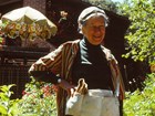 Smiling woman with gardening belt stands in lush garden, wooden home with brick chimney behind her.