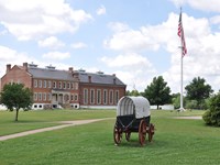 A supply wagon in the grass in front of a large flag pole and the red brick visitor center.