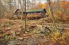 A rustic camp in a wooded area with a wooden bridge, stone steps. Deciduous trees have fall foliage.