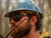 A bearded firefighter in a blue helmet and yellow shirt holds a radio with antenna near his mouth.