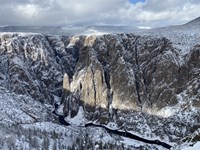 Black Canyon walls and rim have snow, sky has clouds. Dark river is at the bottom.