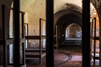 Wooden bunkbeds in a brick building as seen through iron prison bars.