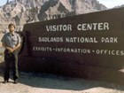 A man with a brown beard and park ranger uniform with the Badlands National Park Visitor Center sign