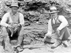 A historic photo of two men seated on rocks with a fossil plant slab propped by the man on the left.