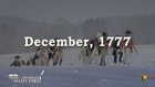 Soldiers in Revolutionary War uniforms walk through a snowy field carrying canvas and wooden posts.