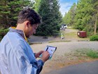 A man writing on a campsite report card. Bear box and evergreen trees are in the background.
