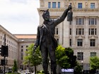 Dark bronze statue of a man in a feathered cap holding a lantern and a pistol