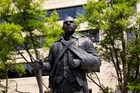 Dark bronze statue of a man with his left arm in a sling and holding a rifle in his right hand