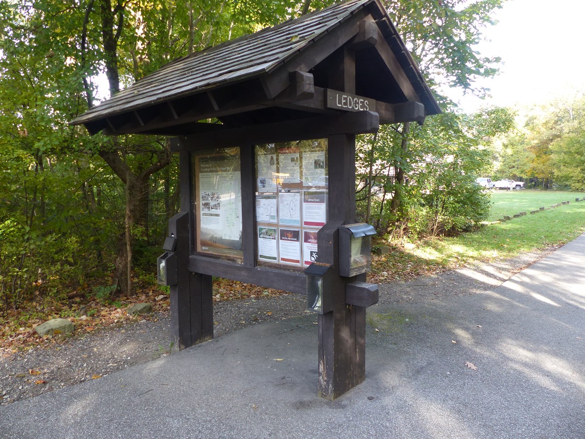 A two-sided bulletin board along a paved road with a Ledges sign below the peaked roof.